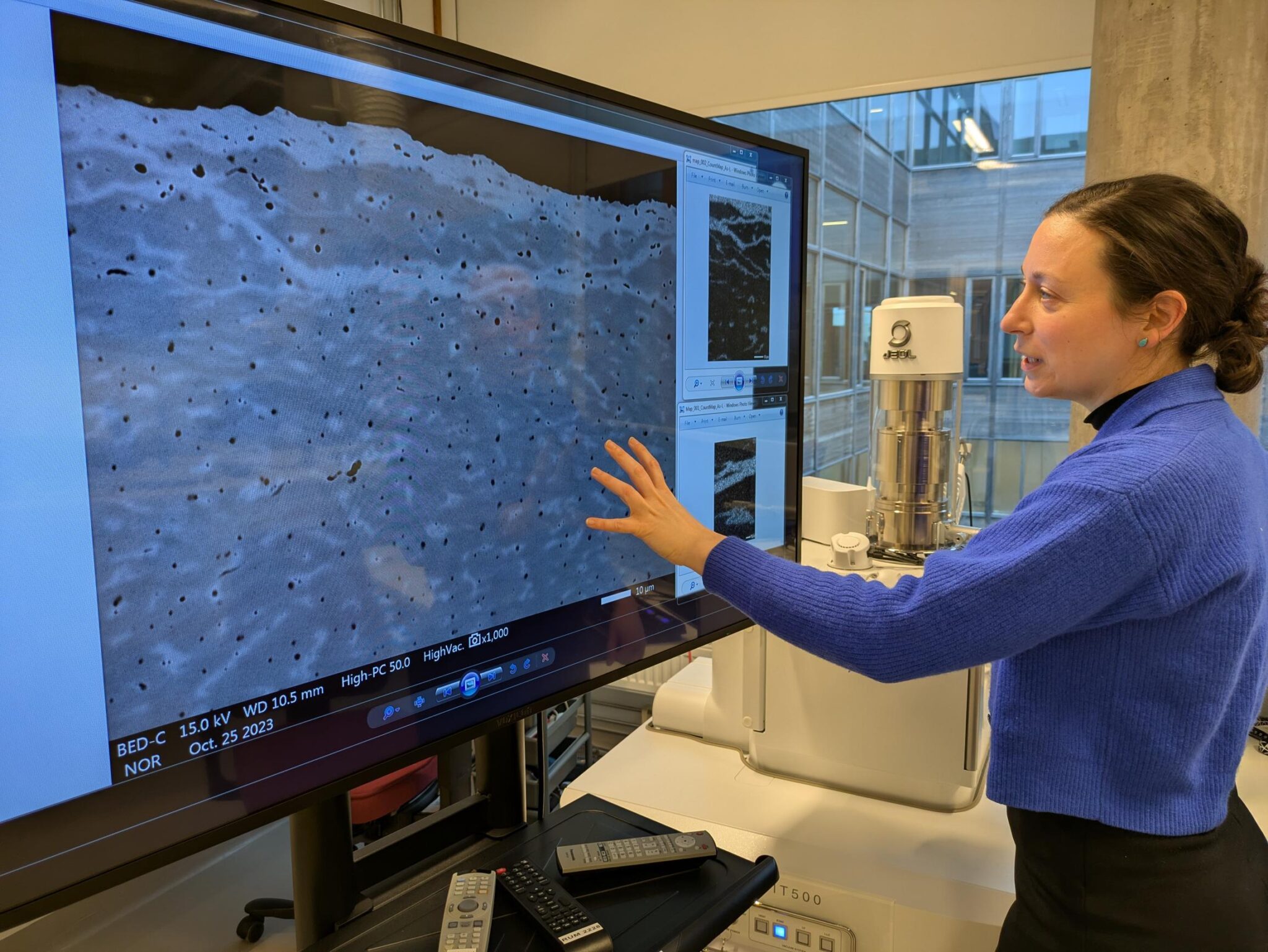 En person i blå kofta står vid en stor skärm och pekar på en förstoring av en materialytas struktur i ett labb med avancerad mikroskoputrustning.