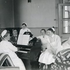 Ett svartvitt foto från 1918 och föreställer leende kvinnor och män i sjukhuskläder. En kvinna spelar piano och de andra sitter runt henne.