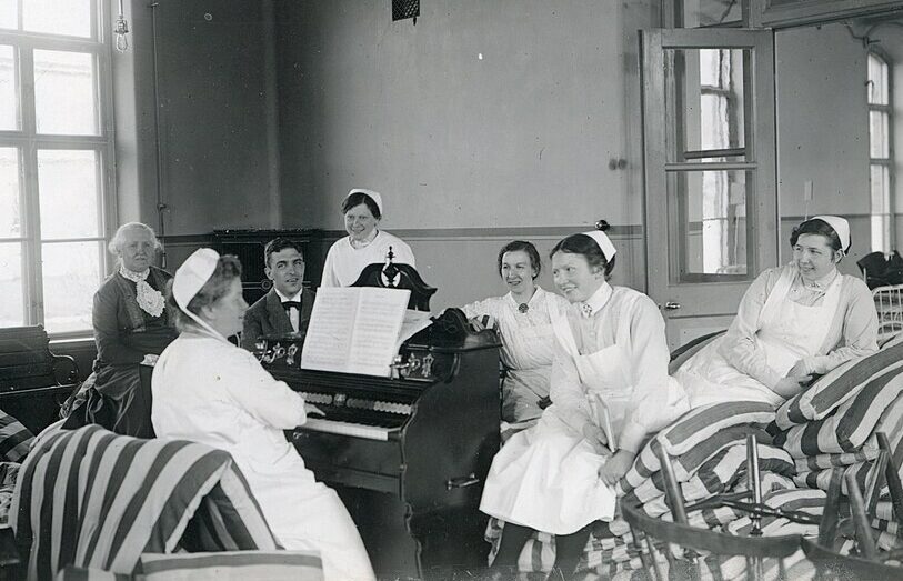Ett svartvitt foto från 1918 och föreställer leende kvinnor och män i sjukhuskläder. En kvinna spelar piano och de andra sitter runt henne.