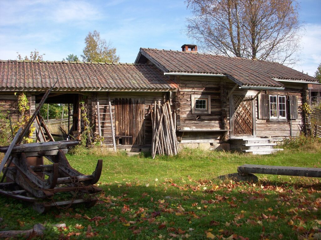 Ett äldre grånat timmerhus med tegeltak står i höstljus, omgivet av gräs, löv och gamla träredskap i förgrunden.