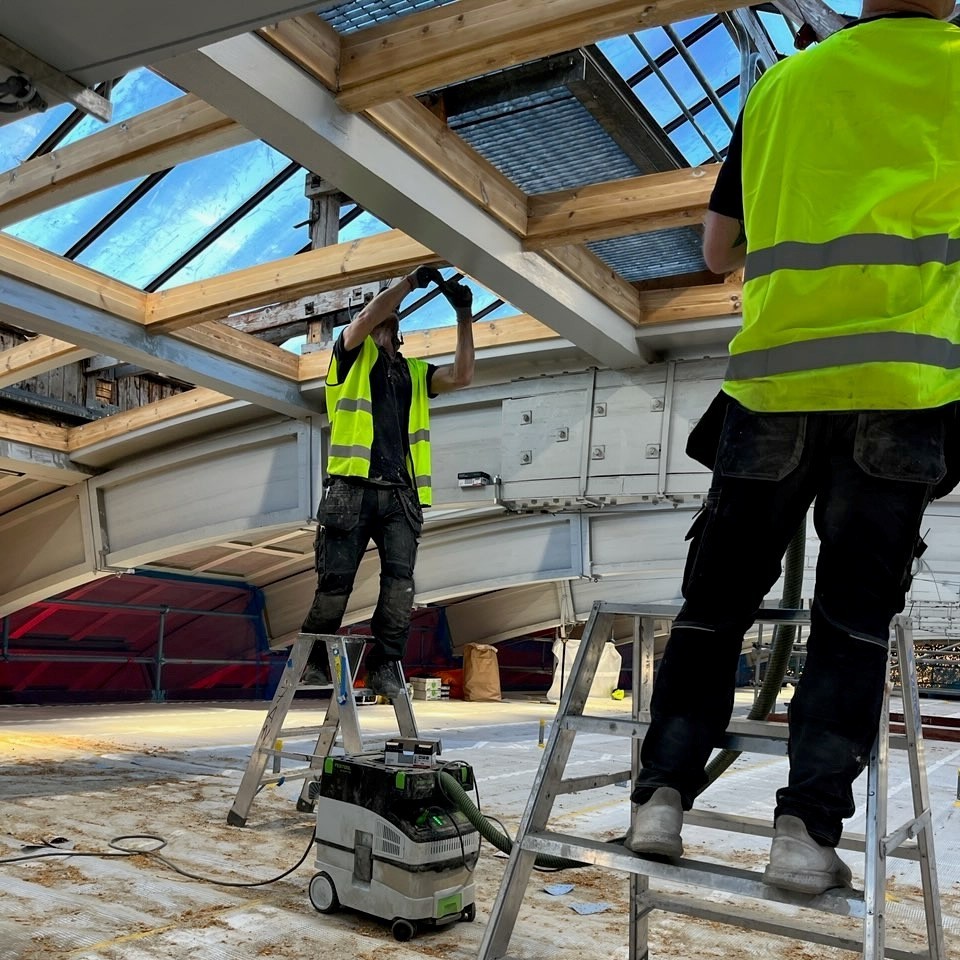 Två byggnadsarbetare i gula reflexvästar står på stegar och arbetar med träkonstruktionen under ett glastak på en renoveringsplats.