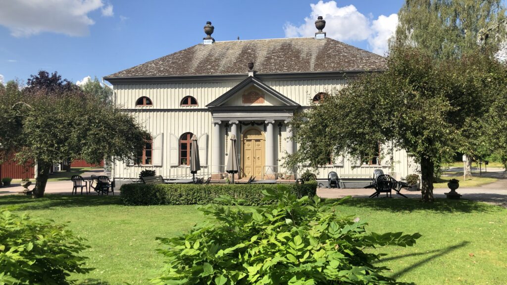 Större byggnad med ljus träfasad och veranda med kolonner omgiven av gröna träd och en solbelyst trädgård framför.