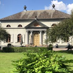 Större byggnad med ljus träfasad och veranda med kolonner omgiven av gröna träd och en solbelyst trädgård framför.