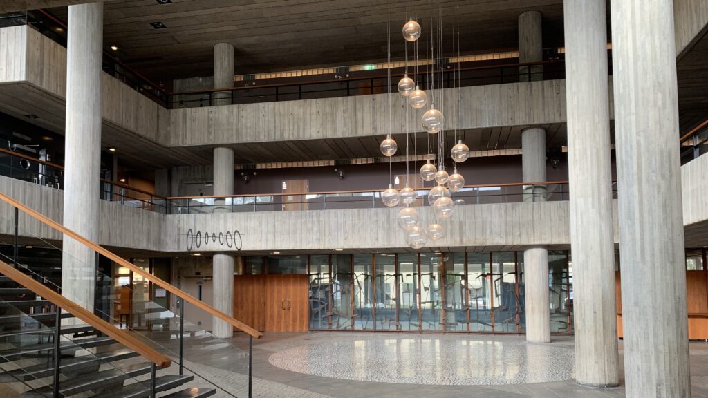 Rymlig entréhall i betong med öppna loftgångar, stora pelare och en klunga hängande glaskulor som belysning.