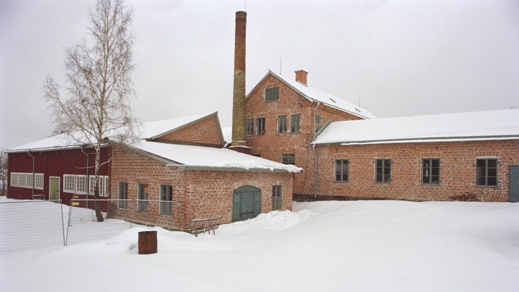 Snötäckt gårdsplan framför äldre industribyggnader i rött tegel med hög skorsten och vinterblek himmel.
