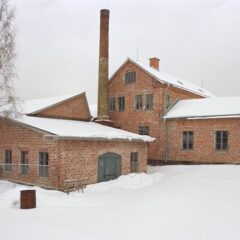 Snötäckt gårdsplan framför äldre industribyggnader i rött tegel med hög skorsten och vinterblek himmel.