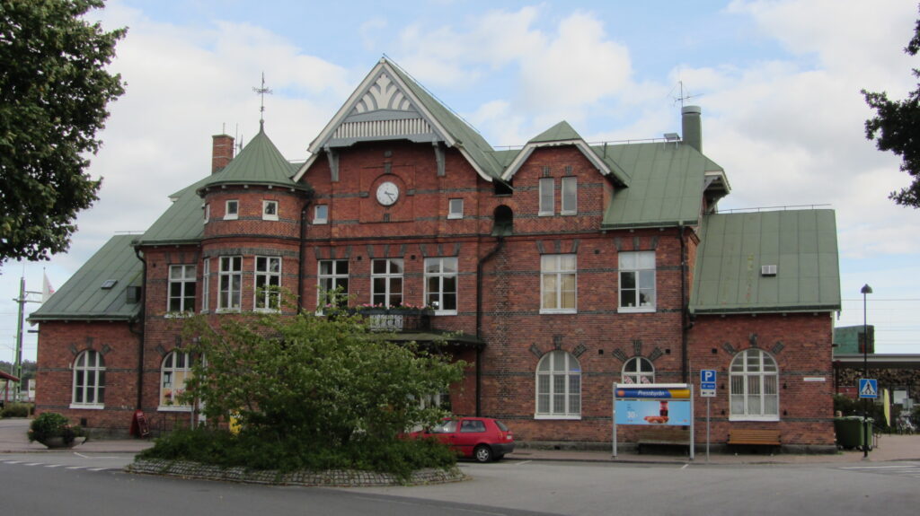 Rött tegelhus i två våningar med grönt sadeltak, rundade och spetsiga fönster samt en klocka på fasaden, sett från gatan framför byggnaden.