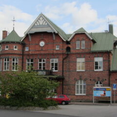 Rött tegelhus i två våningar med grönt sadeltak, rundade och spetsiga fönster samt en klocka på fasaden, sett från gatan framför byggnaden.