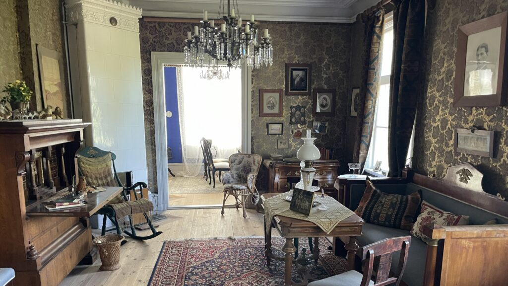 Gammaldags salong med mörka tapeter, antika möbler och kristallkrona, med utsikt mot ett ljusare angränsande rum.