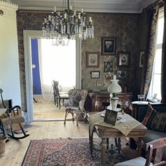 Gammaldags salong med mörka tapeter, antika möbler och kristallkrona, med utsikt mot ett ljusare angränsande rum.