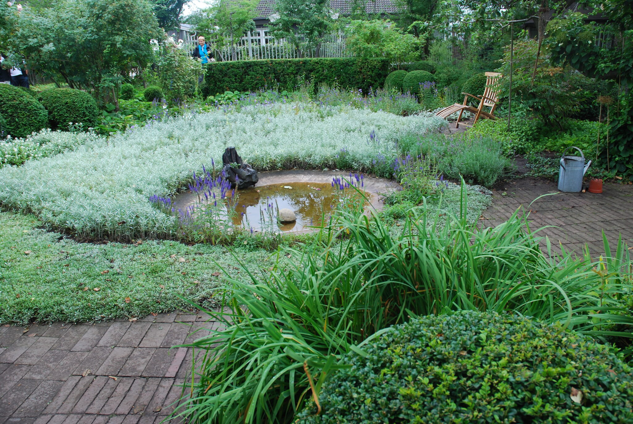 En trädgård med stenlagda gångar och planteringar med rabatter, träd och buskar. I mitten en liten damm.