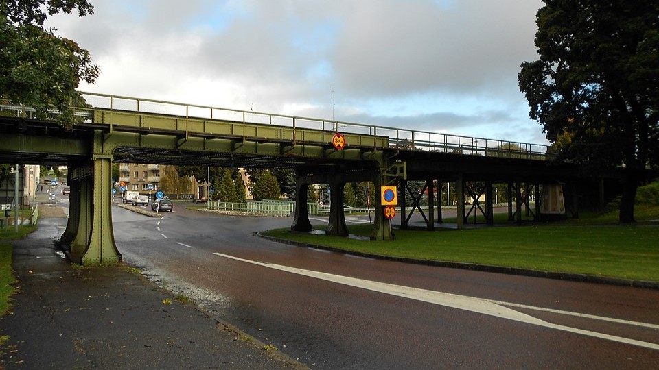 Grönmålad järnvägsviadukt på höga stålben som korsar en bred väg, omgiven av träd och gräs under mulen himmel.