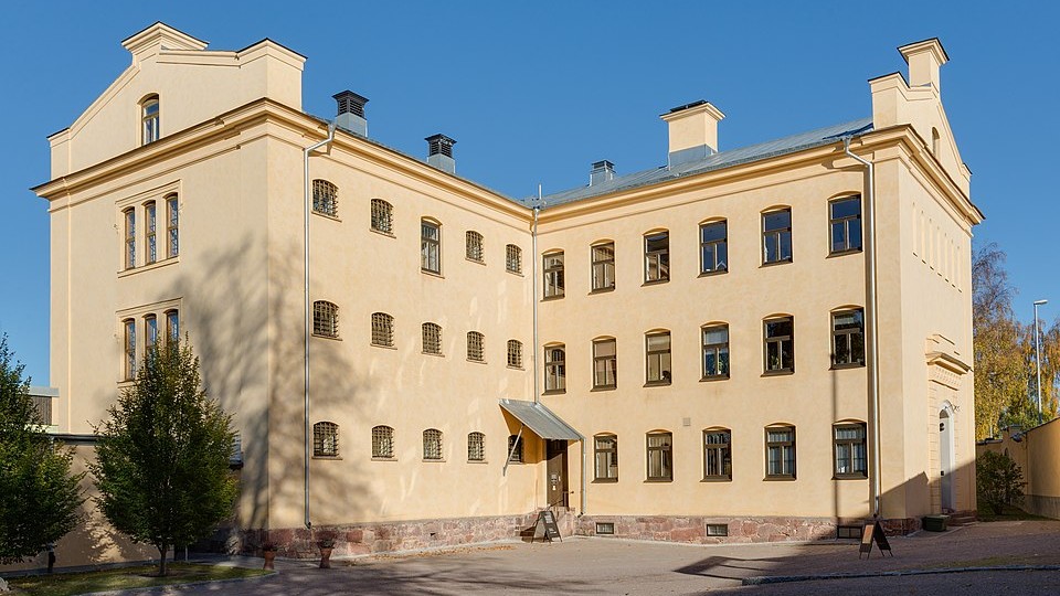 Ljust gul byggnad i tre våningar med höga fönster och spetsade gavlar, belyst av sol mot klar blå himmel.