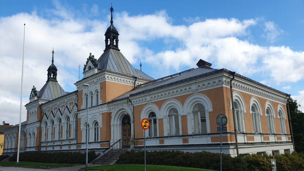 Ljusrosa och vit historisk byggnad med torn och kupoler mot blå himmel, omgiven av buskar och en tom flaggstång i förgrunden.