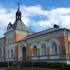 Ljusrosa och vit historisk byggnad med torn och kupoler mot blå himmel, omgiven av buskar och en tom flaggstång i förgrunden.