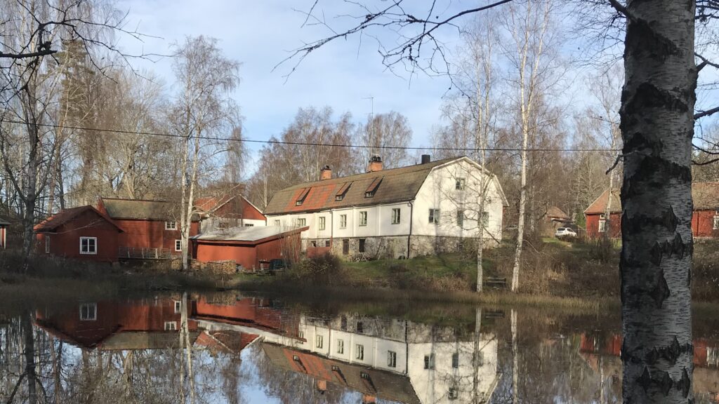 Gårdsbyggnader i rött och vitt speglas i en stilla vattendrag, omgivna av kala björkar i blekt vårljus.