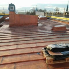 Låglutande koppartak under renovering i eftermiddagssol, med ventilationshuv, låga takkupor och byggmaterial på en pall i förgrunden.