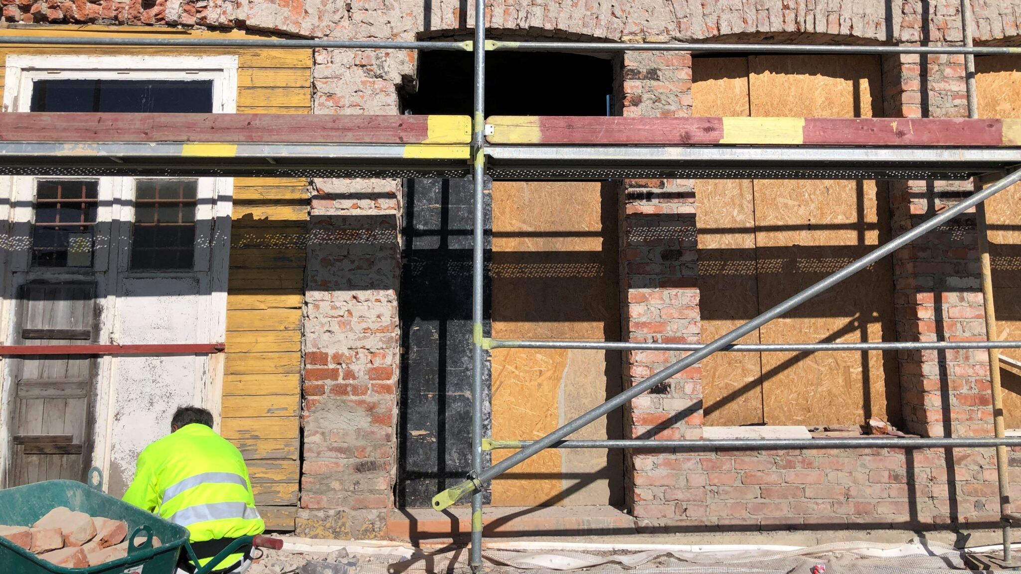 Fasad under renovering med avskalad puts och tegel, byggnadsställning framför och en person i gul varseljacka som arbetar vid en skottkärra i förgrunden.