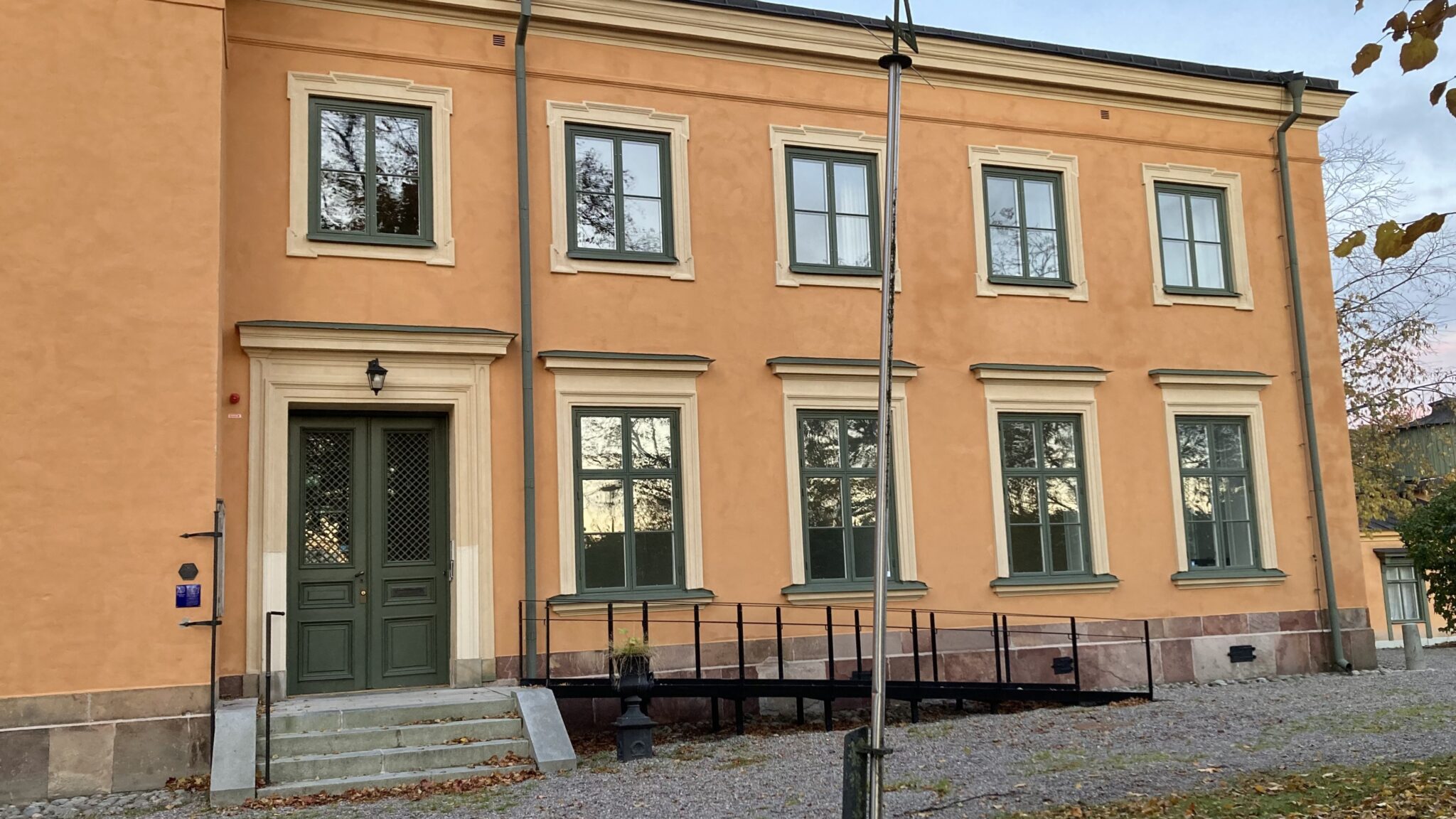 Gulputsad tvåvåningsbyggnad med gröna dubbeldörrar, symmetriskt placerade fönster och en svart ramp framför entrén, omgiven av höstlöv och grusplan.