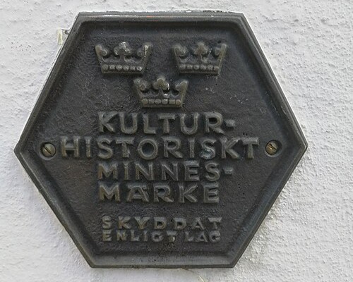Sexkantig mörk metallplakett med tre kronor och texten ”Kulturhistoriskt minnesmärke” monterad på en ljus vägg.