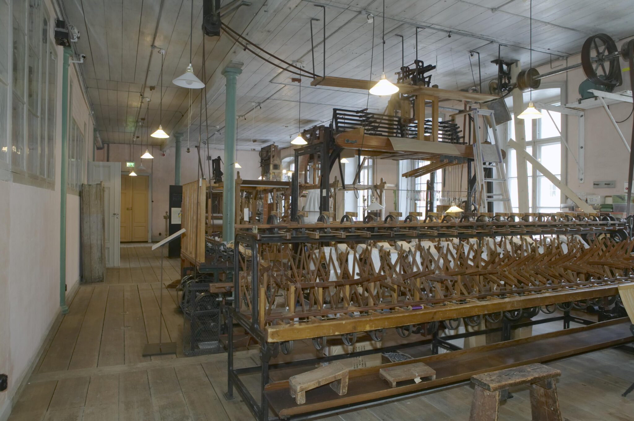 Interiör från en äldre uindustrilokal, med industriella vävstolar i trä som drivs av remdrift.