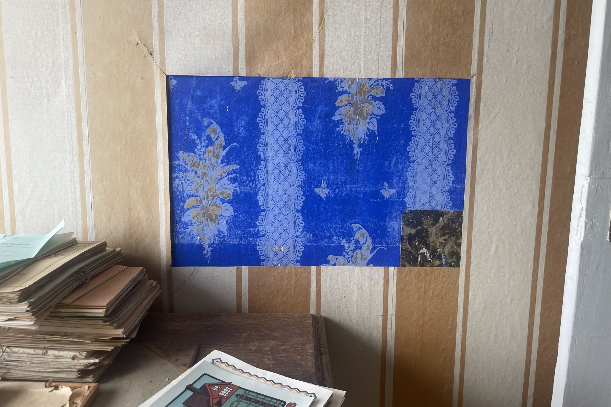 Utskuret parti i en randig tapet som visar ett äldre, blått tapetlager med ljusa mönster och en liten mörkare bit i nedre hörnet; pappershögar ligger på ett bord intill.