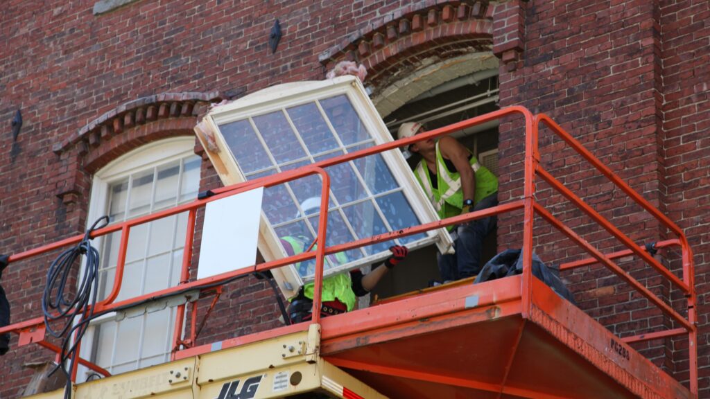 Två personer på en orange skylift lyfter in ett stort fönster i en röd tegelväggs öppning.