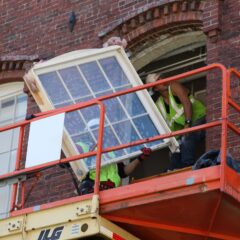 Två personer på en orange skylift lyfter in ett stort fönster i en röd tegelväggs öppning.