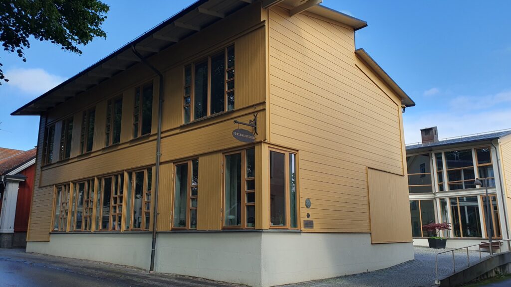 Ett stort gult trähus med många höga fönster och putsad sockel, beläget vid en lugn gata under blå himmel.