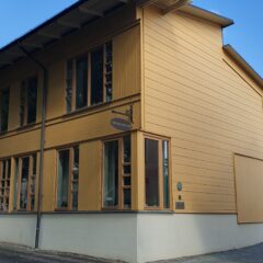 Ett stort gult trähus med många höga fönster och putsad sockel, beläget vid en lugn gata under blå himmel.