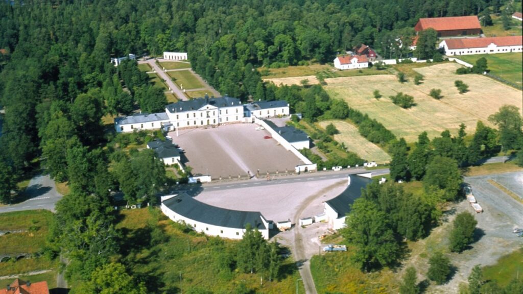 Flygbild över en herrgårdsanläggning omgiven av skog och åkermark, med huvudbyggnad, två flyglar och en stor öppen gårdsplan framför.