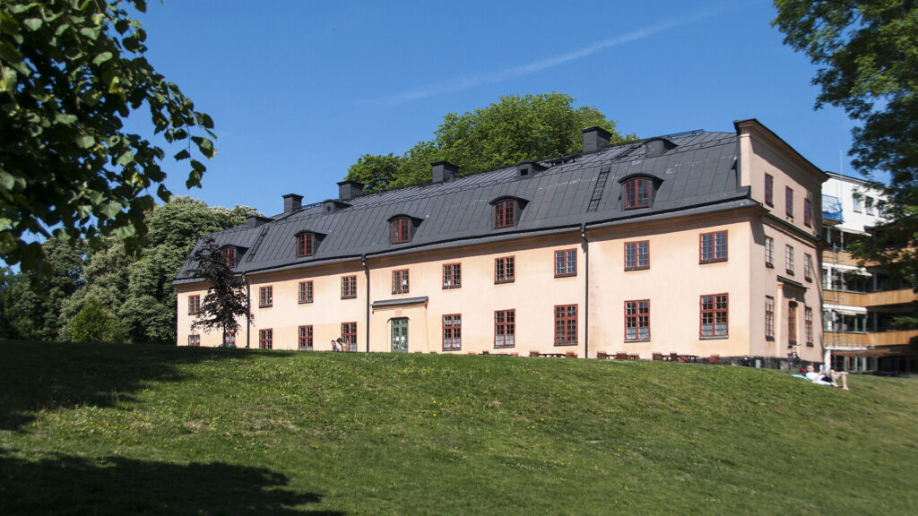 En ljusrosa, tvåvåningsbyggnad med svart tak står på en grön sluttning, omgiven av träd under en klarblå himmel.