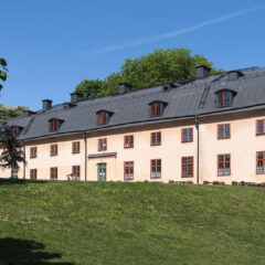 En ljusrosa, tvåvåningsbyggnad med svart tak står på en grön sluttning, omgiven av träd under en klarblå himmel.