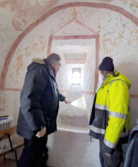 Två personer står i ett valvformat rum med målade väggar och tittar ut genom en djup fönsternisch.