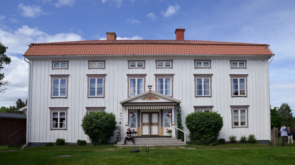 En stor vit träbyggnad i två våningar med rött tegeltak, symmetriska fönsterrader och en utsmyckad entré, omgiven av gräsmatta och buskar.