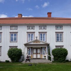 En stor vit träbyggnad i två våningar med rött tegeltak, symmetriska fönsterrader och en utsmyckad entré, omgiven av gräsmatta och buskar.