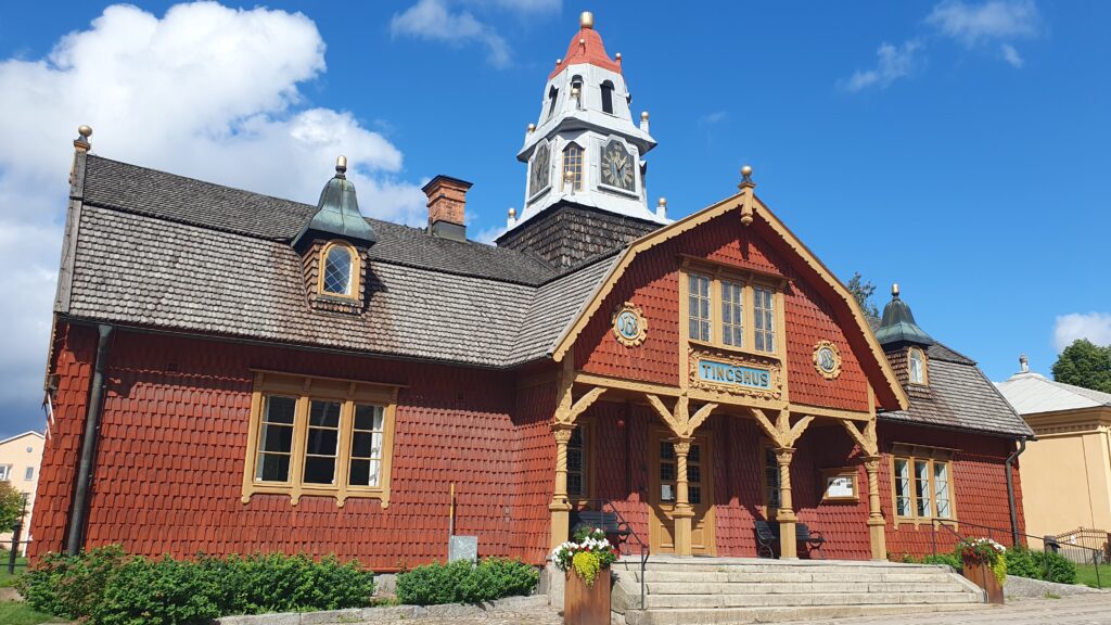 en röd träbyggnad med dekorativa snickerier och en centralt placerad klocktornsliknande överbyggnad med rött tak. Byggnaden har ljusa fönsteromfattningar, en farstukvist med texten ”Tingshus” och ligger i dagsljus under blå himmel med moln.