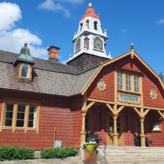 en röd träbyggnad med dekorativa snickerier och en centralt placerad klocktornsliknande överbyggnad med rött tak. Byggnaden har ljusa fönsteromfattningar, en farstukvist med texten ”Tingshus” och ligger i dagsljus under blå himmel med moln.