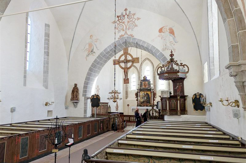 Ett ljust kyrkorum med målade valv, träbänkar och ett stort träkrucifix hängande framför altaret.