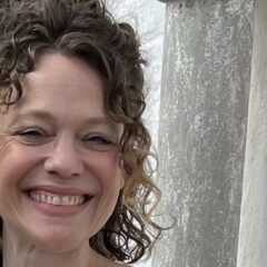 Kvinna med mörkt lockigt uppsatt hår står framför en kalkstensport.