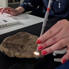 En hand med rött nagellack för ett litet mikroskop över en grå sten med blå fläckar.