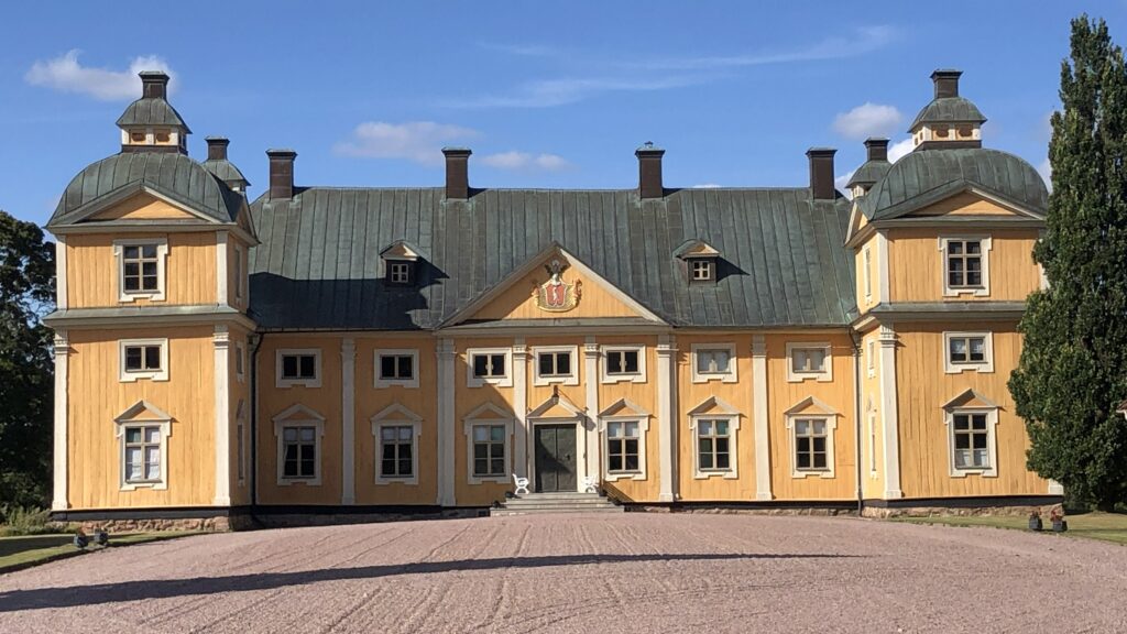 Gult slottsliknande trähus med grönt tak och två tornlika gavlar, sett från grusad gårdsplan under klar blå himmel.