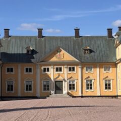 Gult slottsliknande trähus med grönt tak och två tornlika gavlar, sett från grusad gårdsplan under klar blå himmel.