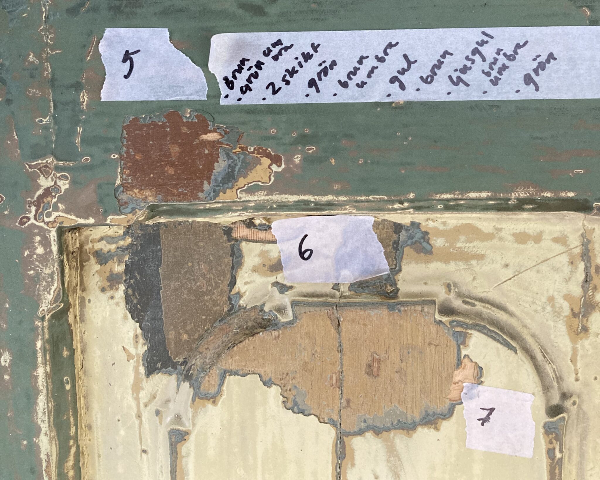 En närbild av en gammal grönmålad dörr där färglagren skrapats fram och märkts med små lappar med siffrorna 5, 6 och 7.