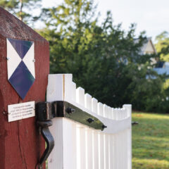 En blåvit skylt sitter på grindstolpen vid en uppställd välkomnade vit trägrind som leder in till ett äldre gult trähus.