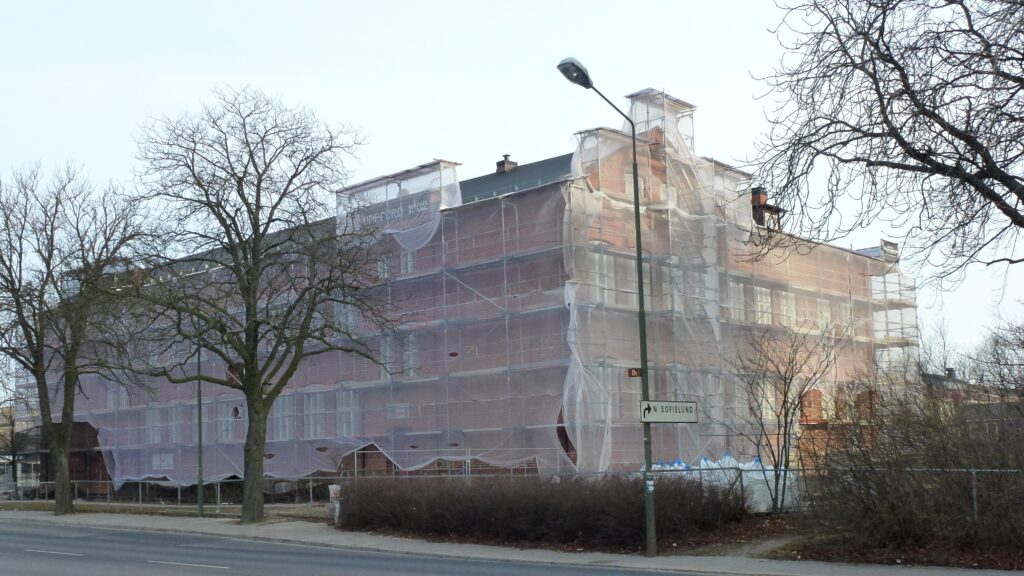 Stort tegelhus helt inhäjt och täckt av byggnadsställningar och skyddsnät längs en gata med nakna träd i förgrunden.
