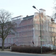 Stort tegelhus helt inhäjt och täckt av byggnadsställningar och skyddsnät längs en gata med nakna träd i förgrunden.