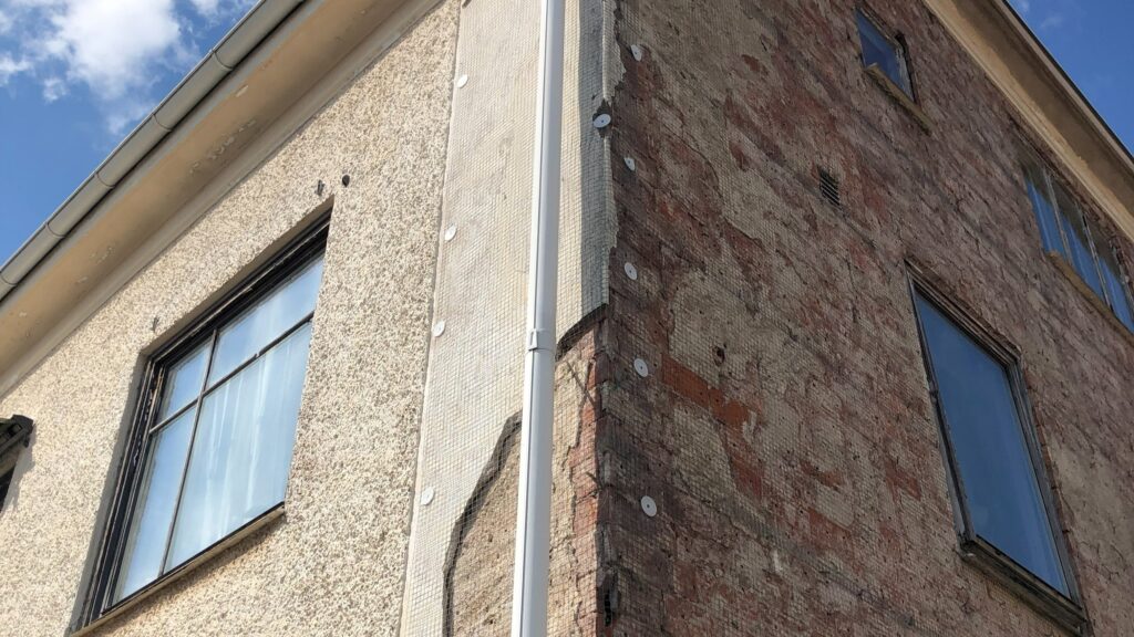 Del av en husfasad där ena sidan har beige puts och den andra visar avskalad röd tegelvägg, med höga fönster och en vit stupränna mitt emellan.