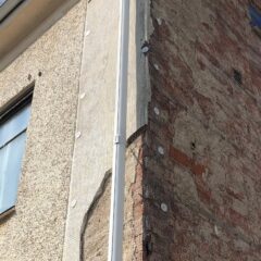 Del av en husfasad där ena sidan har beige puts och den andra visar avskalad röd tegelvägg, med höga fönster och en vit stupränna mitt emellan.