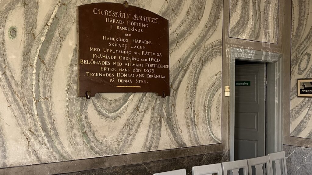 En mönstrad stenvägg med en mörk minnestavla i trä ovanför en rad ljusgrå stolar i ett äldre rum.